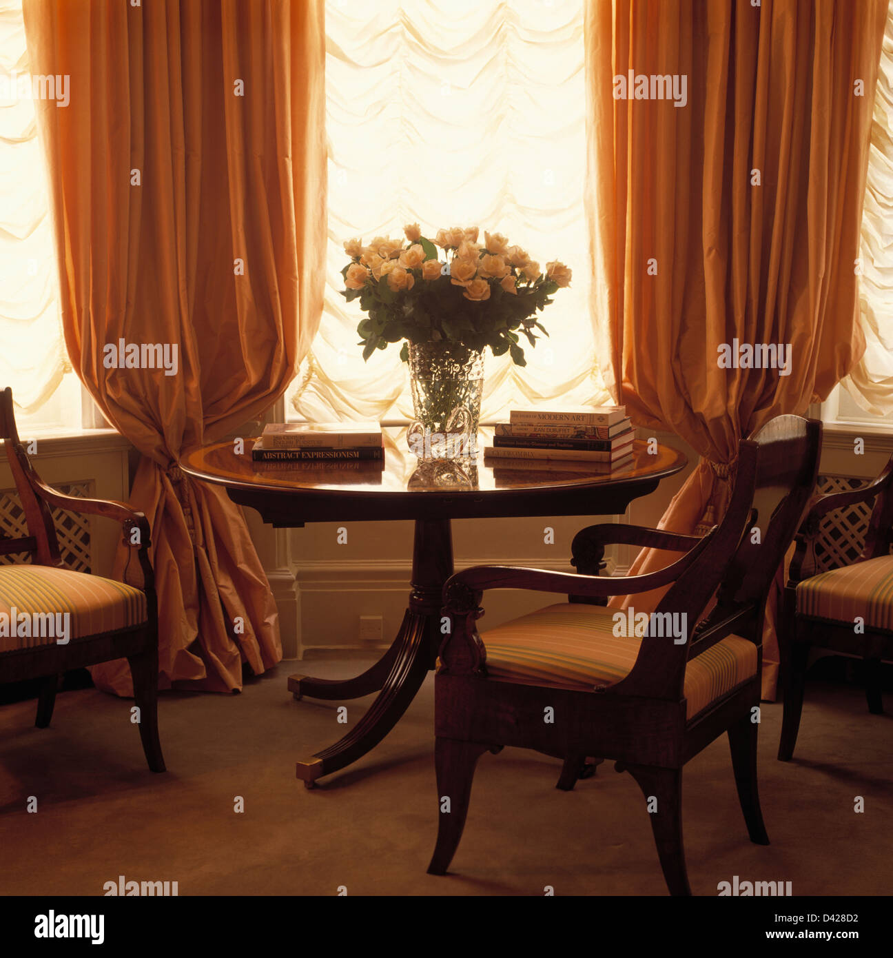 Circular mahogany table and upholstered dining chairs in front of window with ruched blinds and peach silk curtains Stock Photo