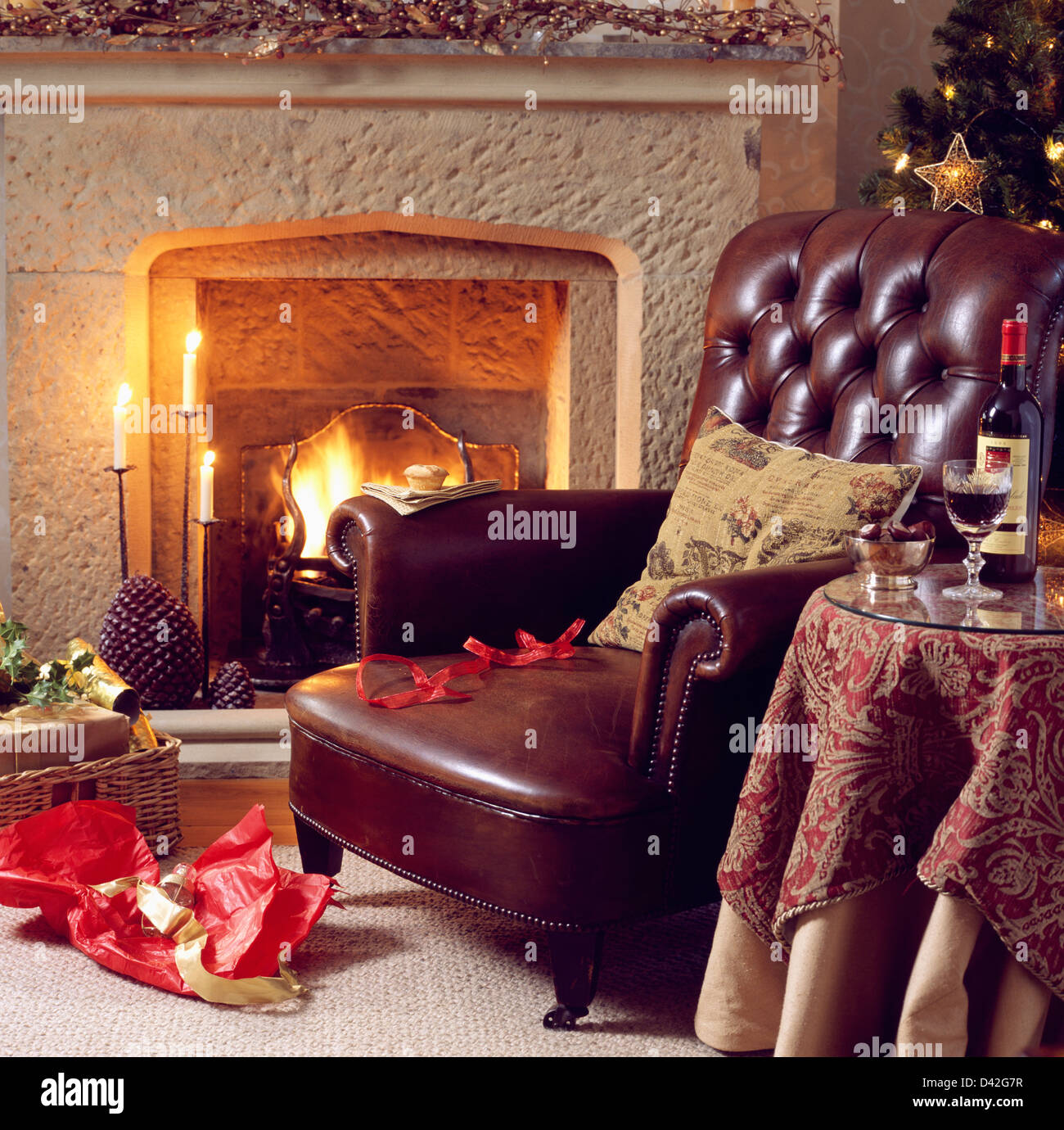 Brown leather sofa beside fireplace with lighted fire in country living room with patterned cloth on small table Stock Photo
