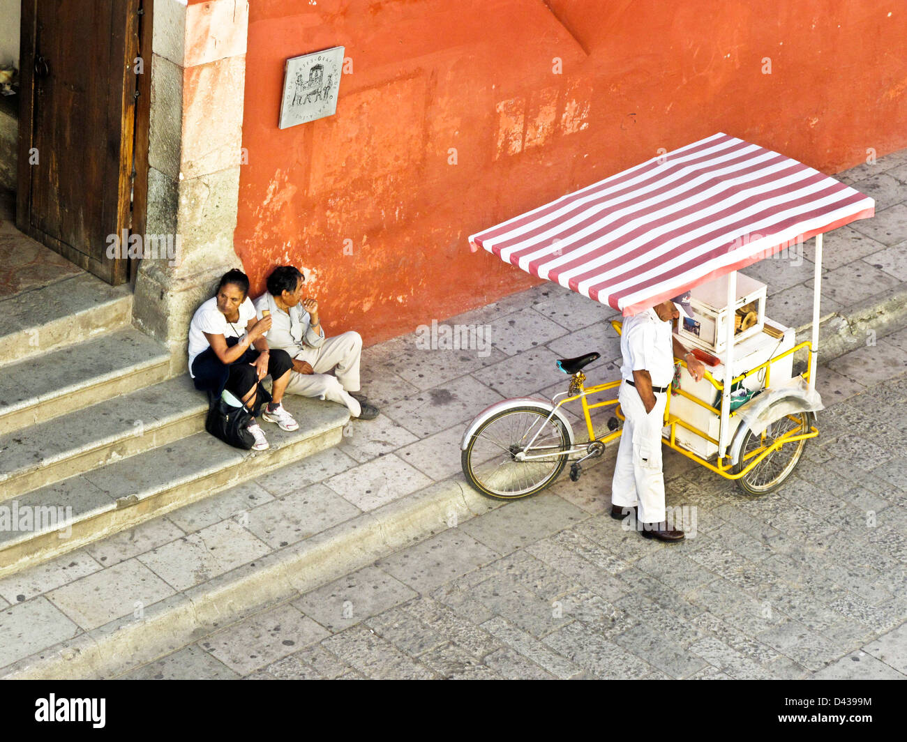 middle aged couple sit on steps of Oaxaca Institute of Graphic Arts with ice cream bought from vendor with cart at curb Mexico Stock Photo