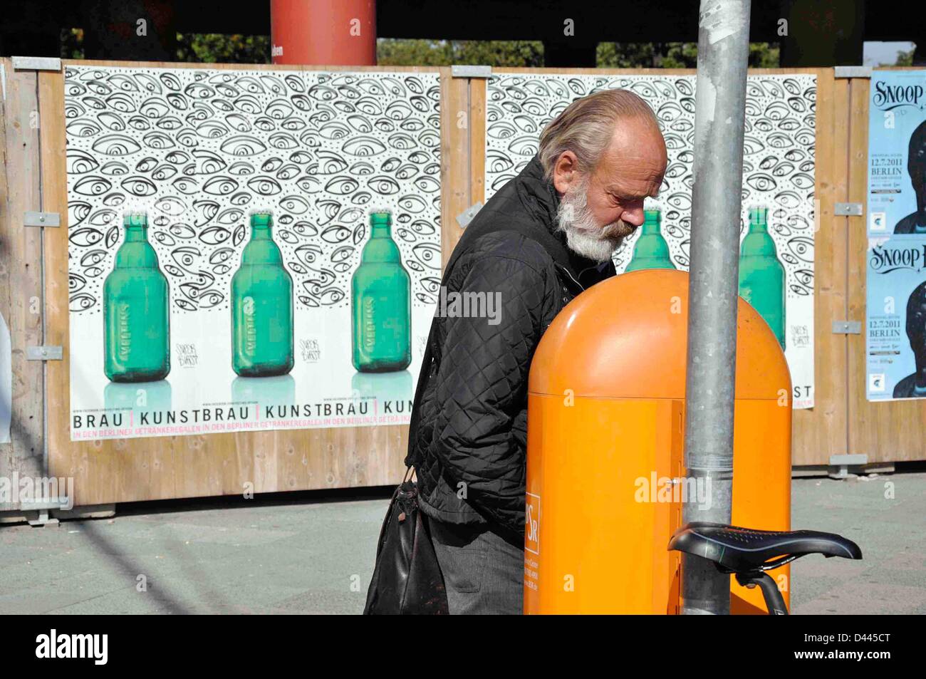An old man searches for deposit bottles and recycle products in an orange trash bin of the waste management company Berliner Stadtreinigungsbetriebe (BSR) in Berlin, Germany, 24 September 2011. In the background, advertisement for beer bottles with modern design by Veltins brewery is pictured. Fotoarchiv für ZeitgeschichteS.Steinach Stock Photo