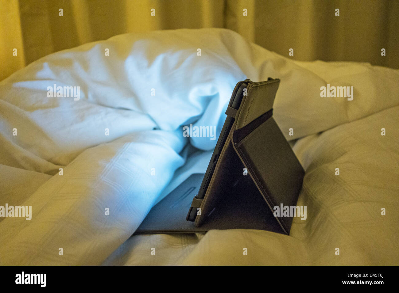 A switched on iPad Mini in its case throwing light onto the bedclothes Stock Photo