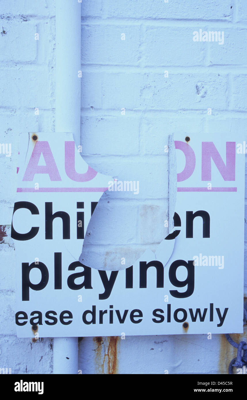 White sign fixed on white painted brick wall with missing pieces stating Caution Children Playing Please Drive Slowly Stock Photo