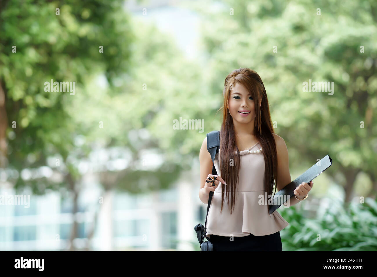 Young woman in business attire, carrying briefcase and holding folder standing outside. Stock Photo