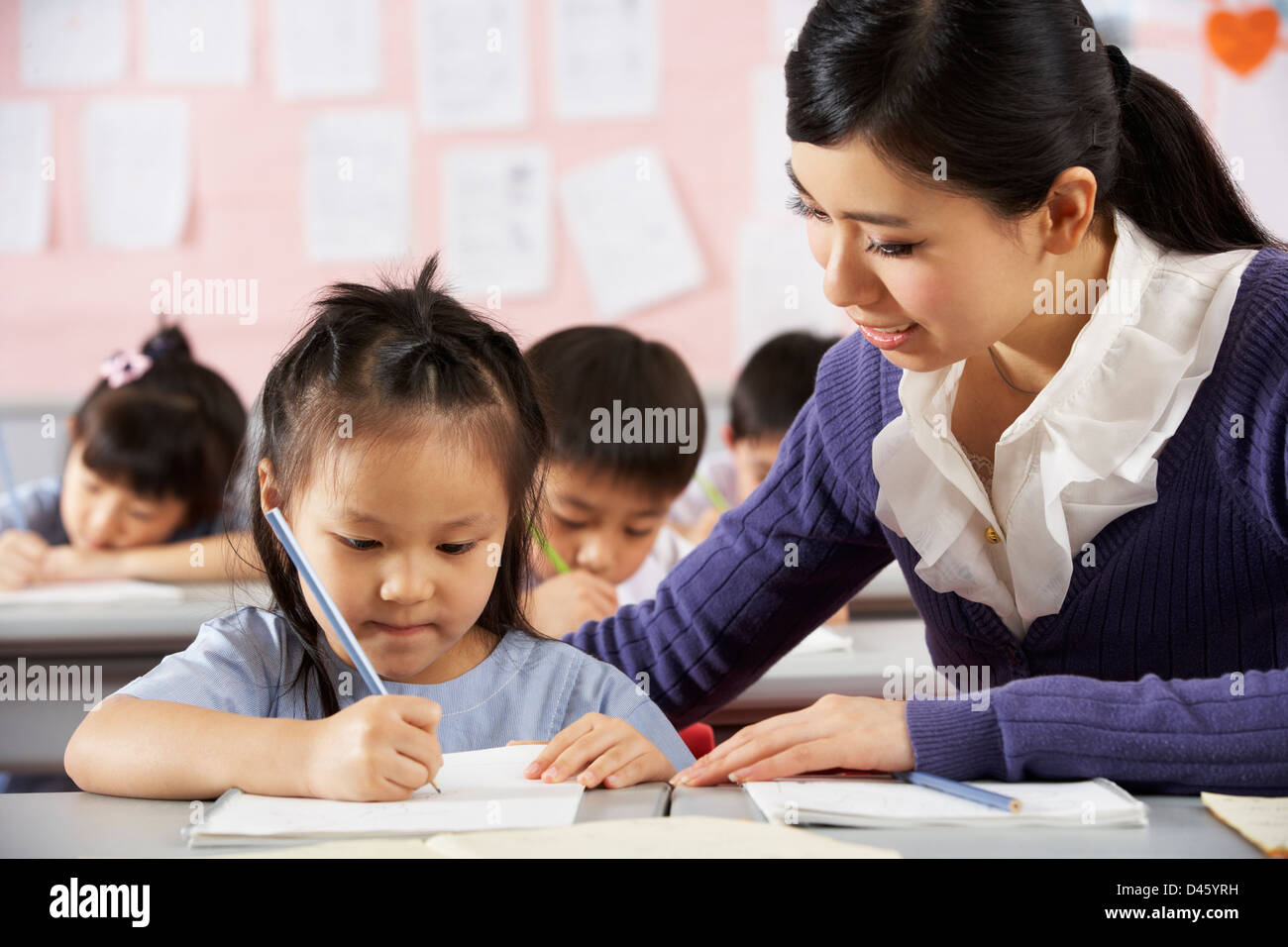 Teacher Helping Student Working At Desk In Chinese School Classroom ...