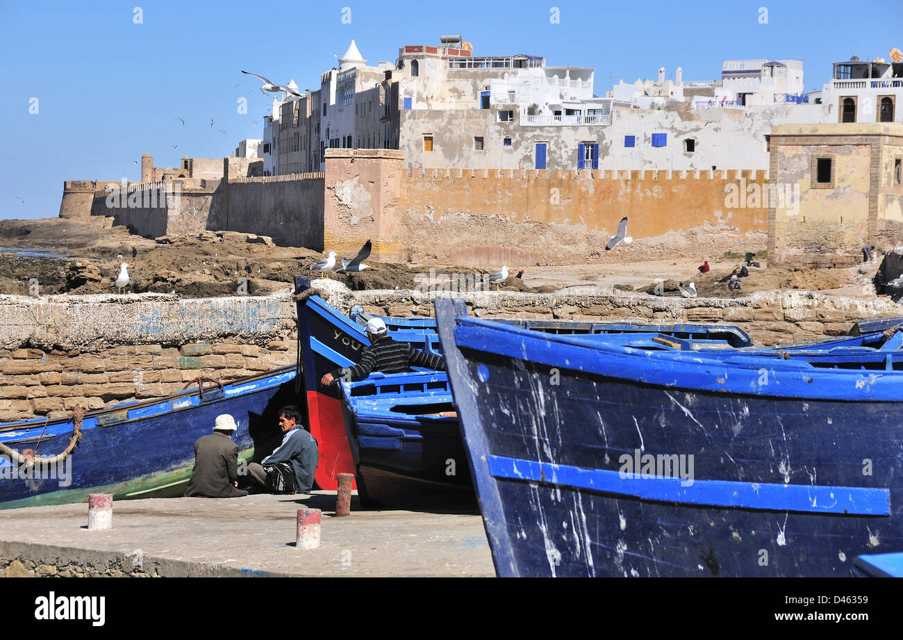 Fisherman chatting beside their fishing boats with seagulls soaring over the  fortified walls of the c-16th coastal town of Essaouira, Morocco Stock Photo