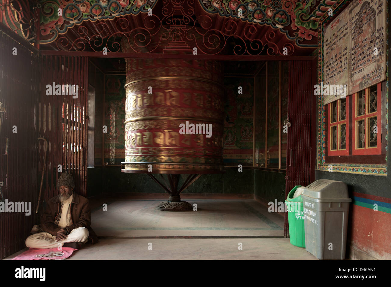 The largest prayer wheel at Boudhanath Stupa and the surrounding temples spins behind an elderly Buddhist worshiper. Stock Photo