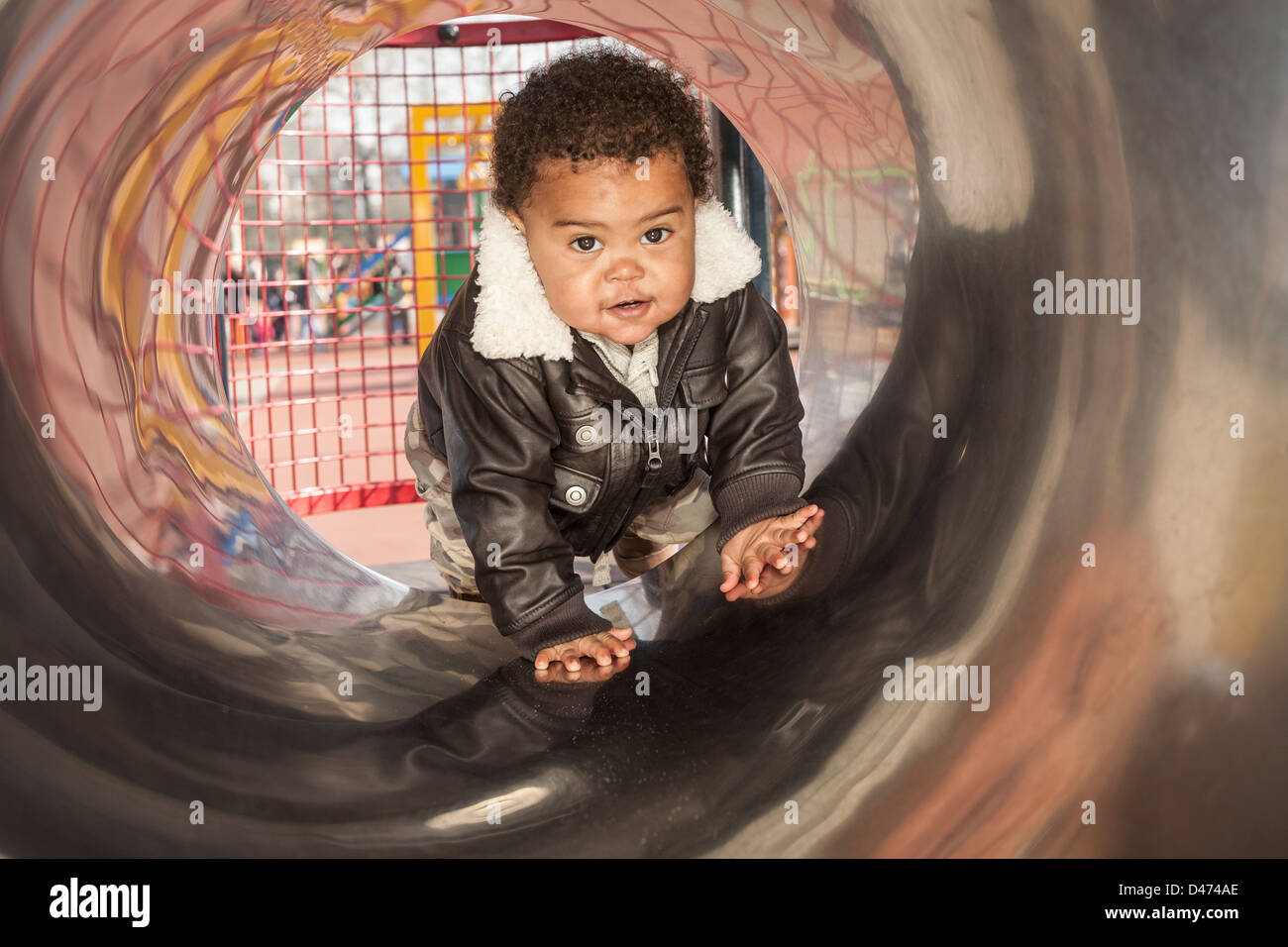 Mixed race toddler baby  playing in metal tunnel at the park wearing aviator jacket crawling on all fours Stock Photo