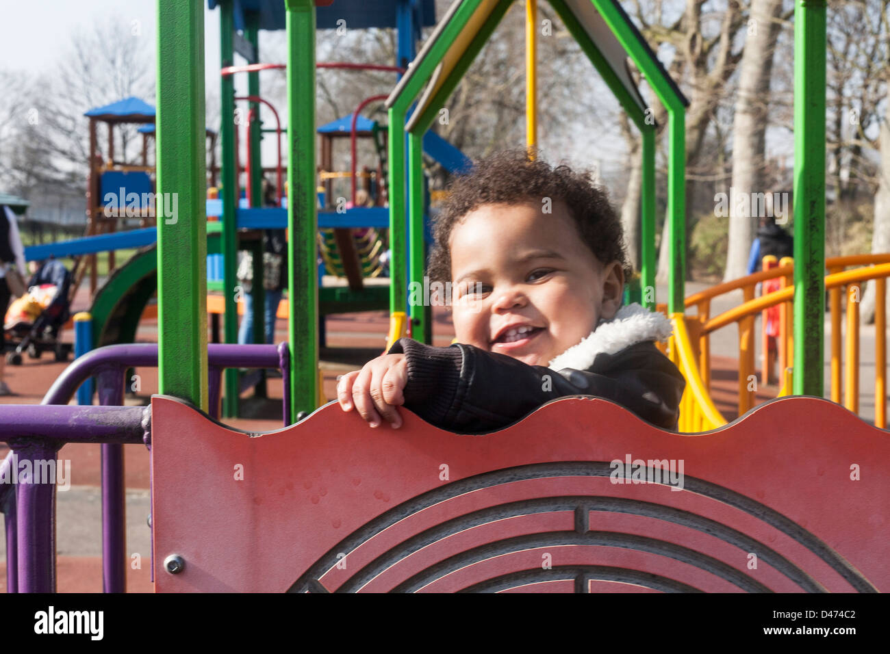 Mixed race toddler baby smiling and playing on climbing frame on a sunny day wearing aviator jacket Stock Photo