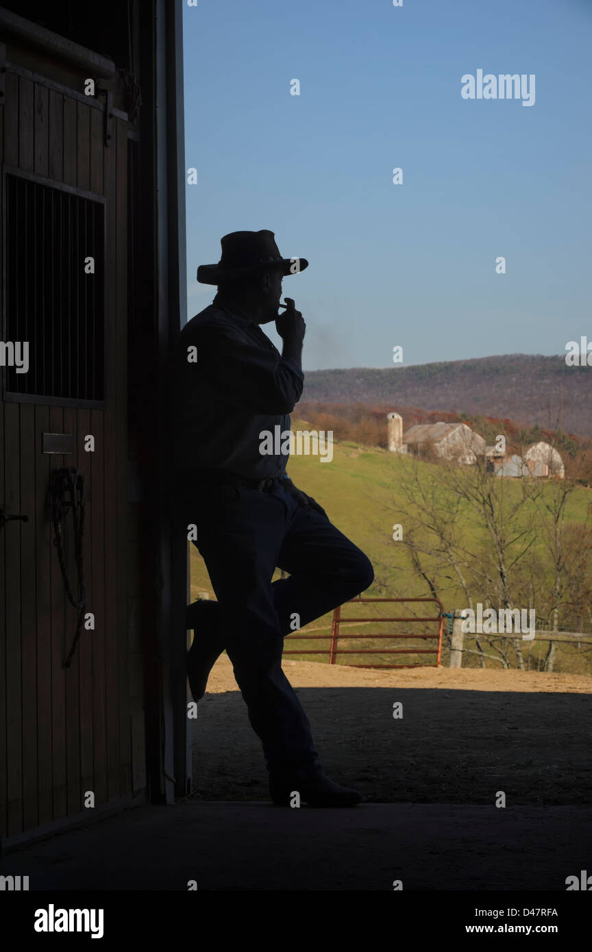Farmer silhouette in open barn door with farm scenery in background, having a smoke in a relaxation pose. Stock Photo