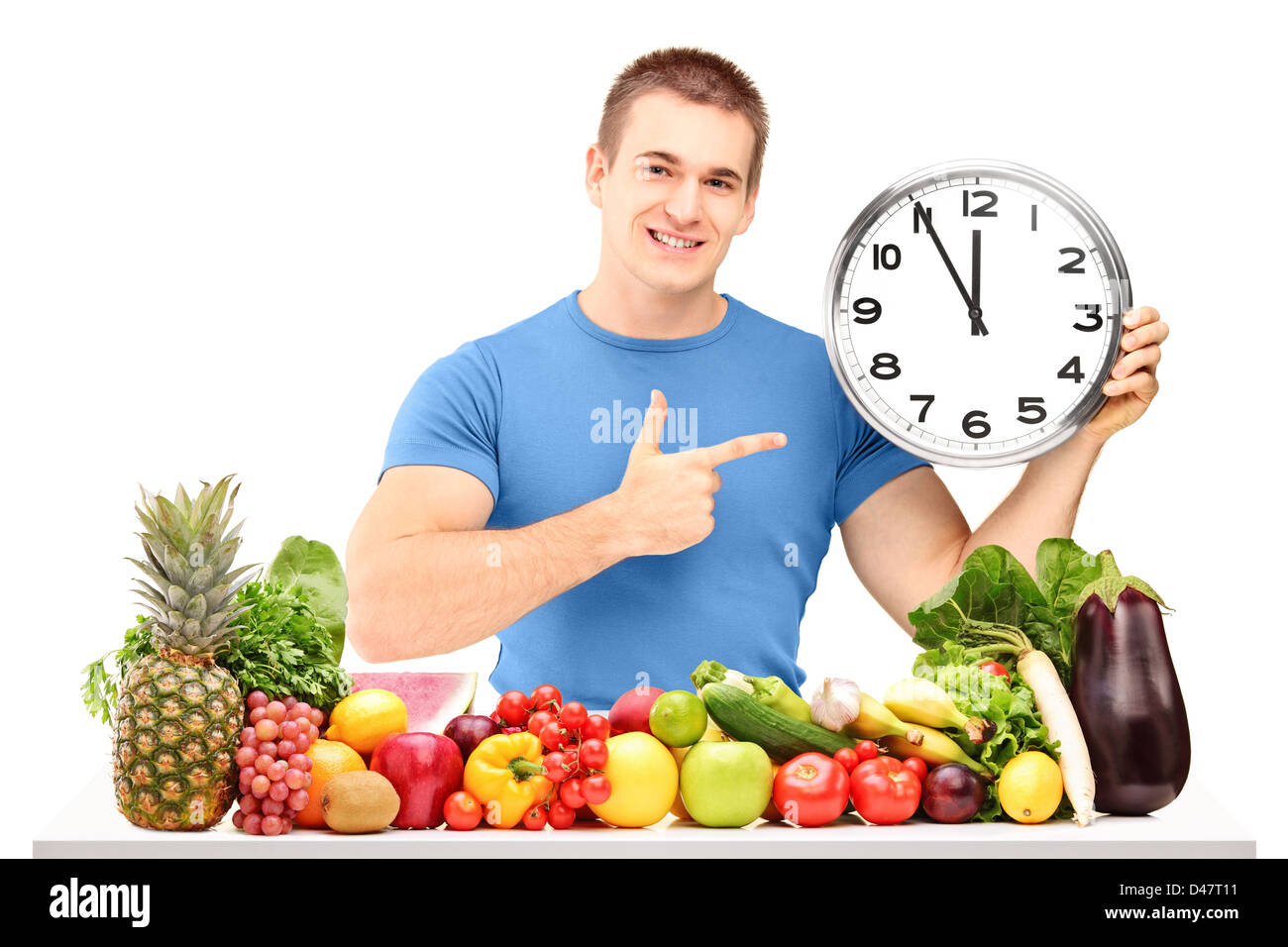 Young man holding a clock and posing with a pile of fruits and vegetables, isolated on white background Stock Photo