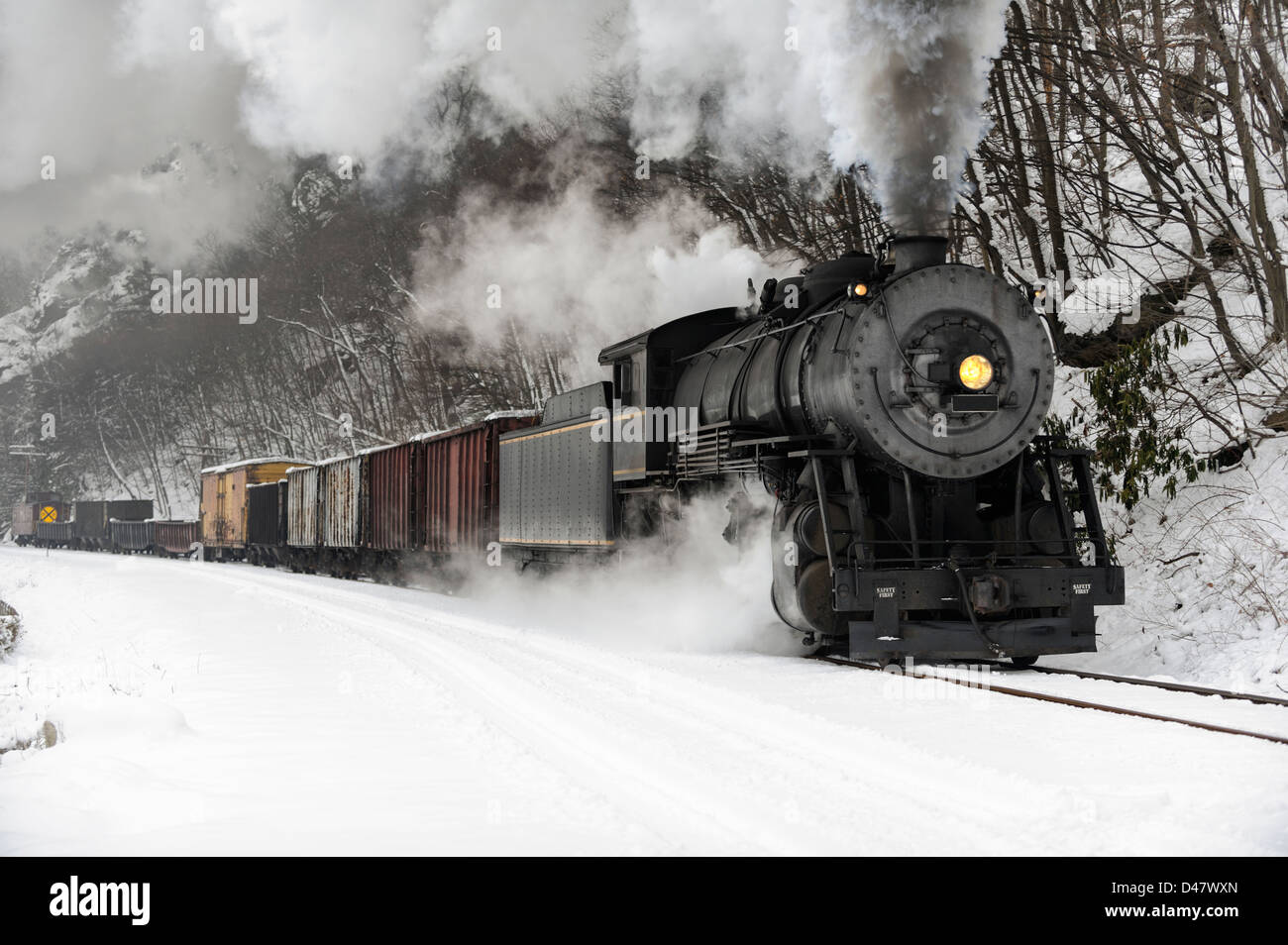 Freight train rounding a curve, through a snowy rock cut, beside a river, a steam locomotive billowing smoke in the cold winter air. Stock Photo