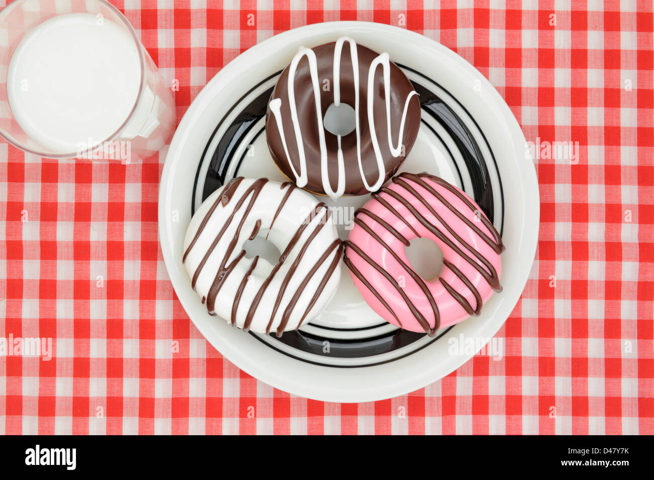 Decorated iced donuts on a white plate, with a glass of milk, on a red-and-white checkered, picnic tablecloth, studio shot taken from above. Stock Photo