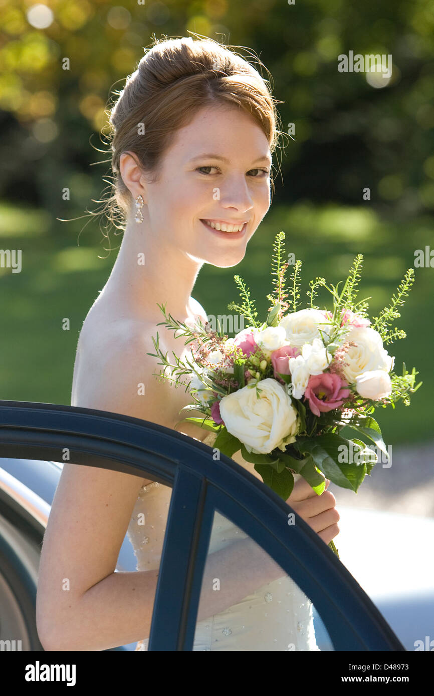 Smiling bride stepping from car side on to the camera in sunshine wearing cream strapless wedding dress holding bouquet Stock Photo
