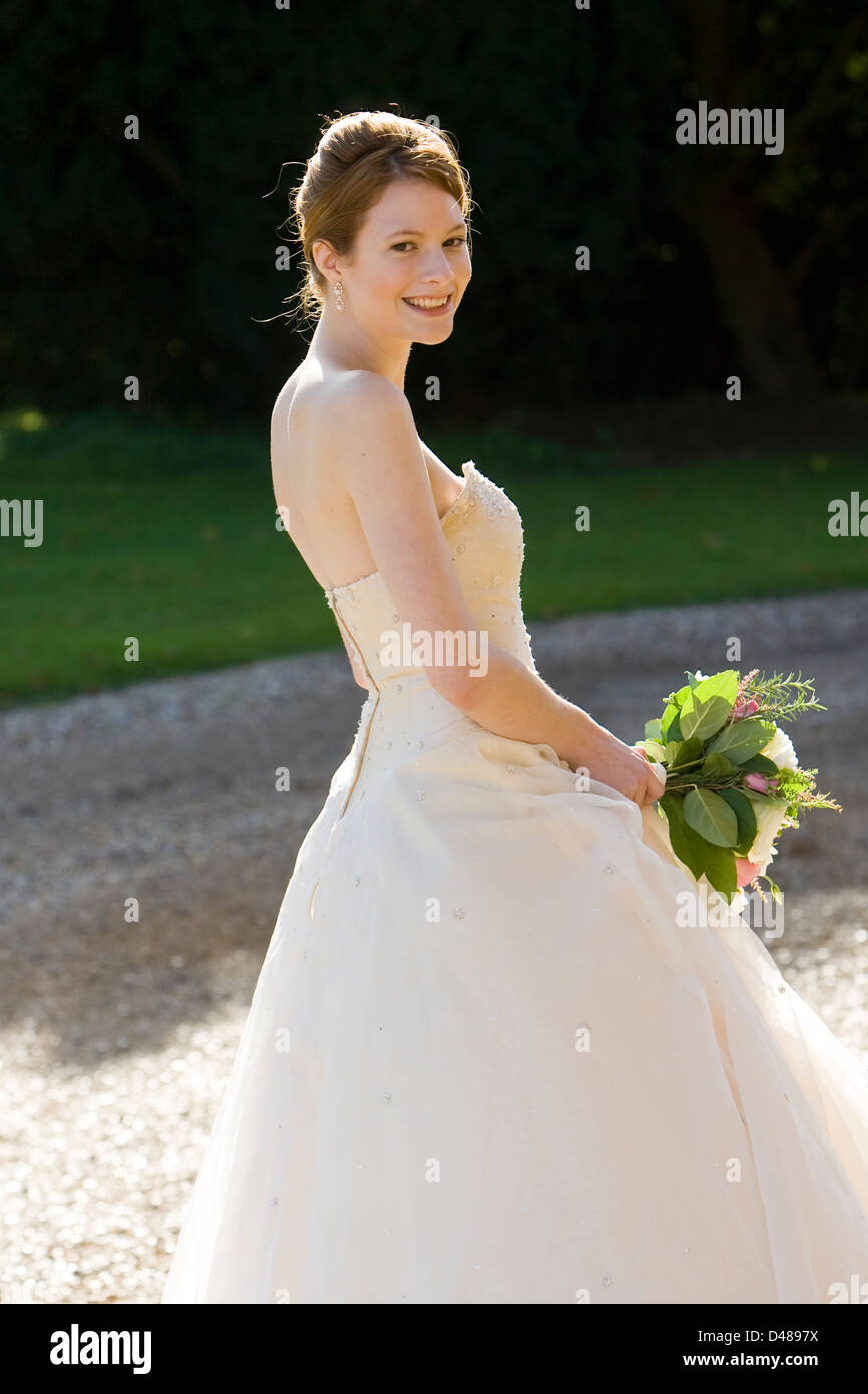 Smiling bride standing side on to the camera in sunshine wearing cream strapless wedding dress holding bouquet Stock Photo