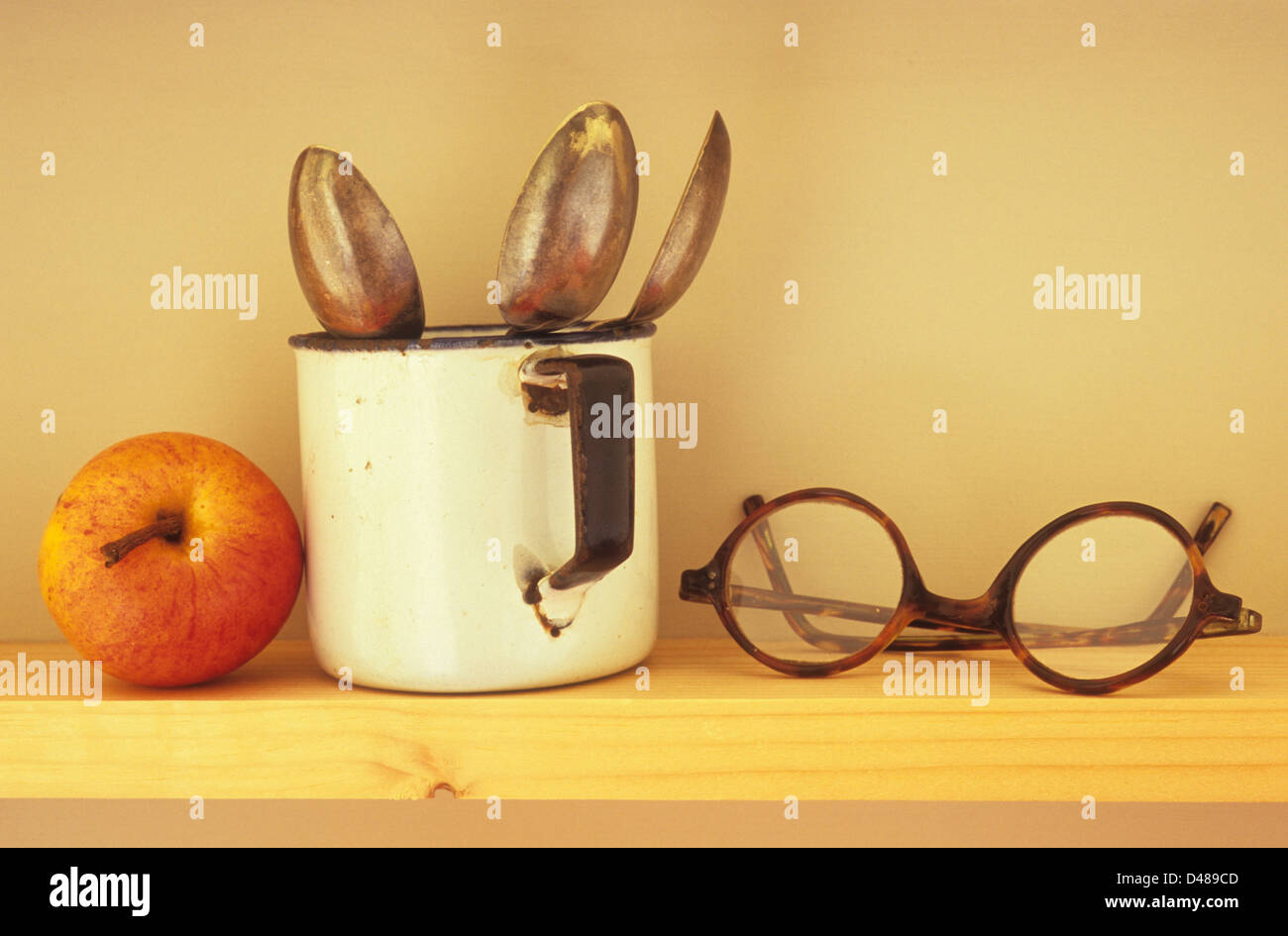 Eye-level view of narrow wooden shelf with apple enamel mug containing three teaspoons and vintage spectacles Stock Photo