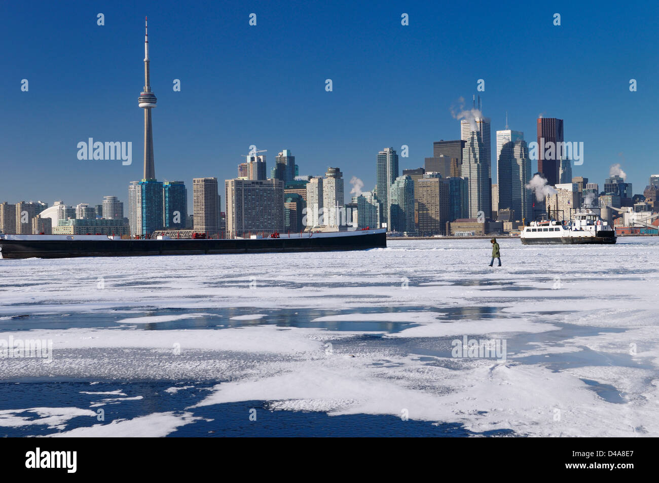 Woman walking on frozen Lake Ontario as the Wards Island Ferry returns to Toronto in winter with city skyline Stock Photo