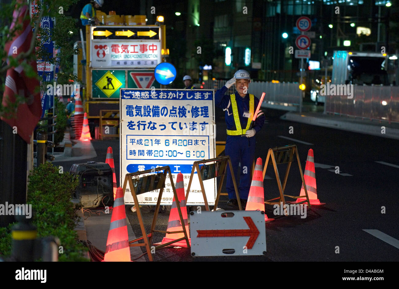 A construction worker flagman salutes as he stands guard over a road repair site on a Tokyo street at night. Stock Photo