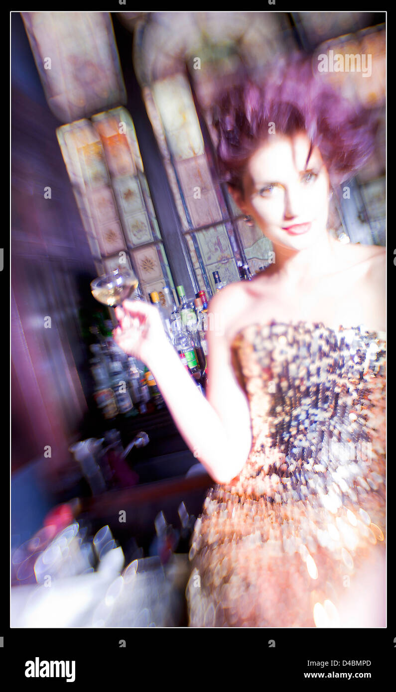A white European female age 25-30 wearing a stylish designer cocktail dress, she's sipping Champagne against a bar in color. Stock Photo