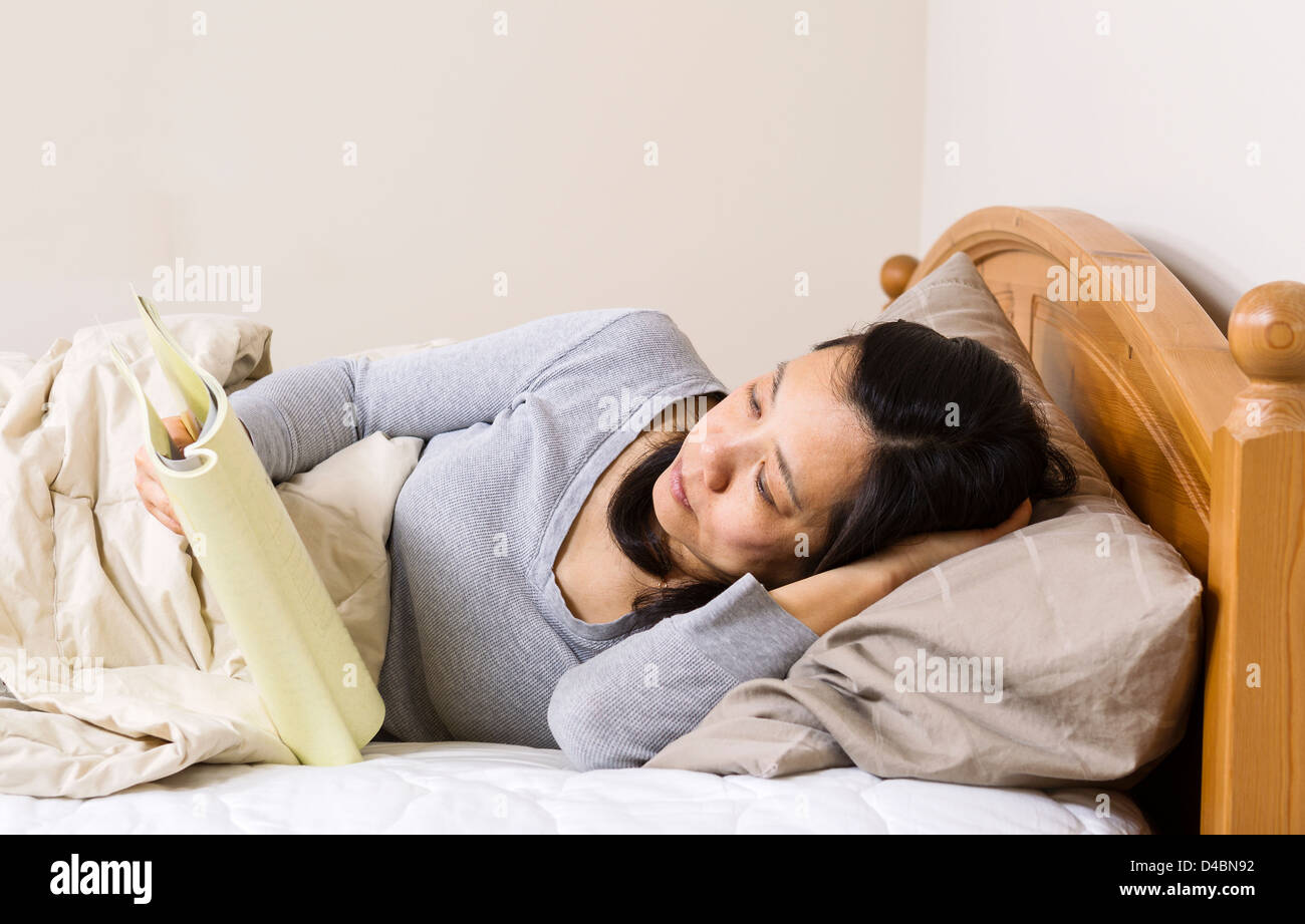 Horizontal photo of mature woman, lying head down in pillow, while reading a book in bed Stock Photo