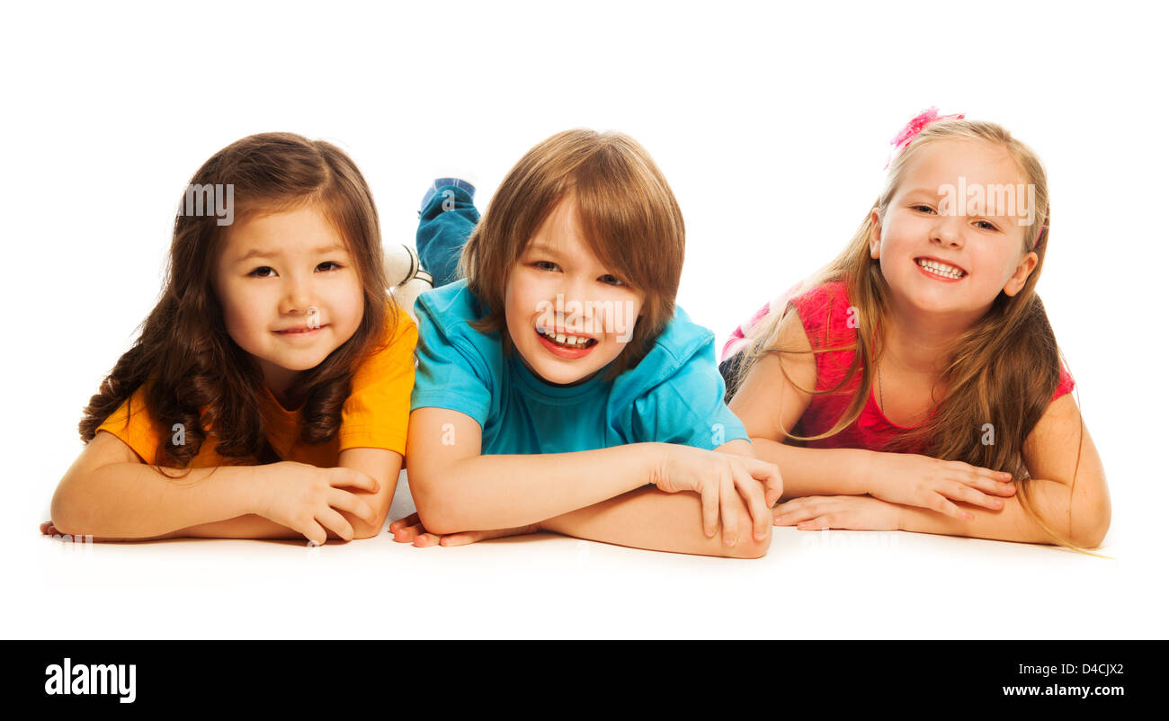 Three happy kids, boy and girls laying on the floor in a line together smiling and laughing, isolated on white Stock Photo