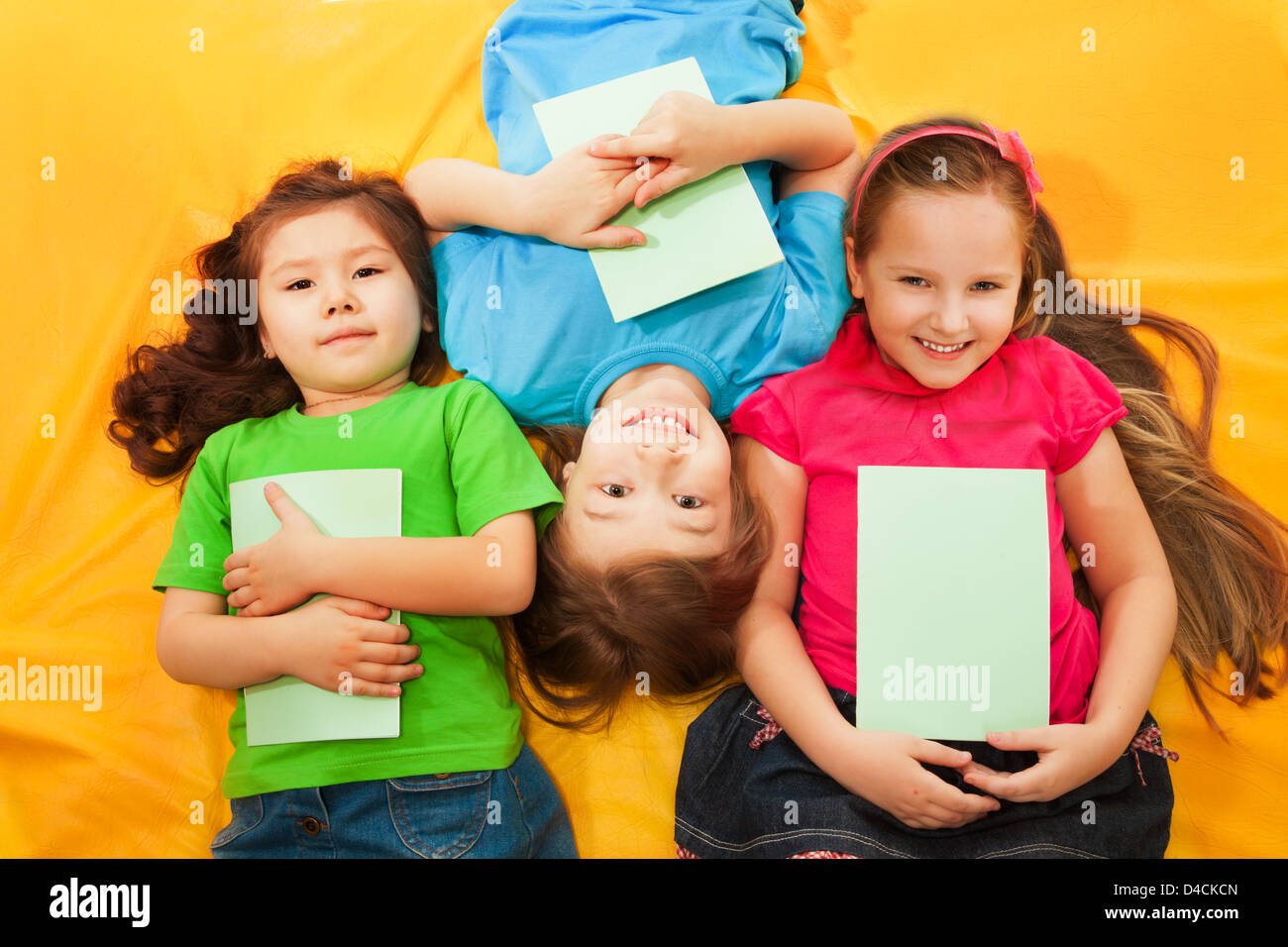 Three little kids, boys and girls, laying with books on the yellow mattress, smiling, with long hair, Stock Photo