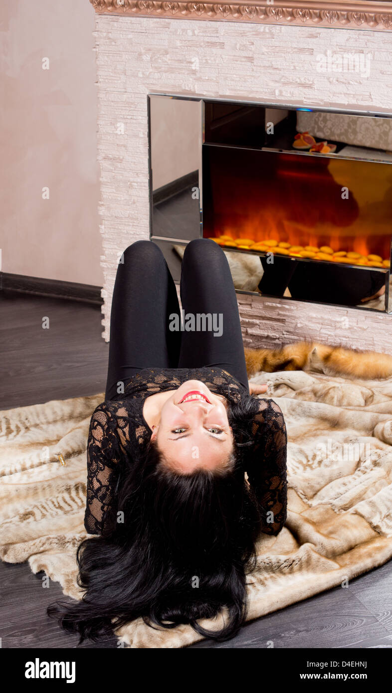 Playful beautiful woman lying on her back in front of a blazing fire on a fur coat looking mischievously up at the camera Stock Photo
