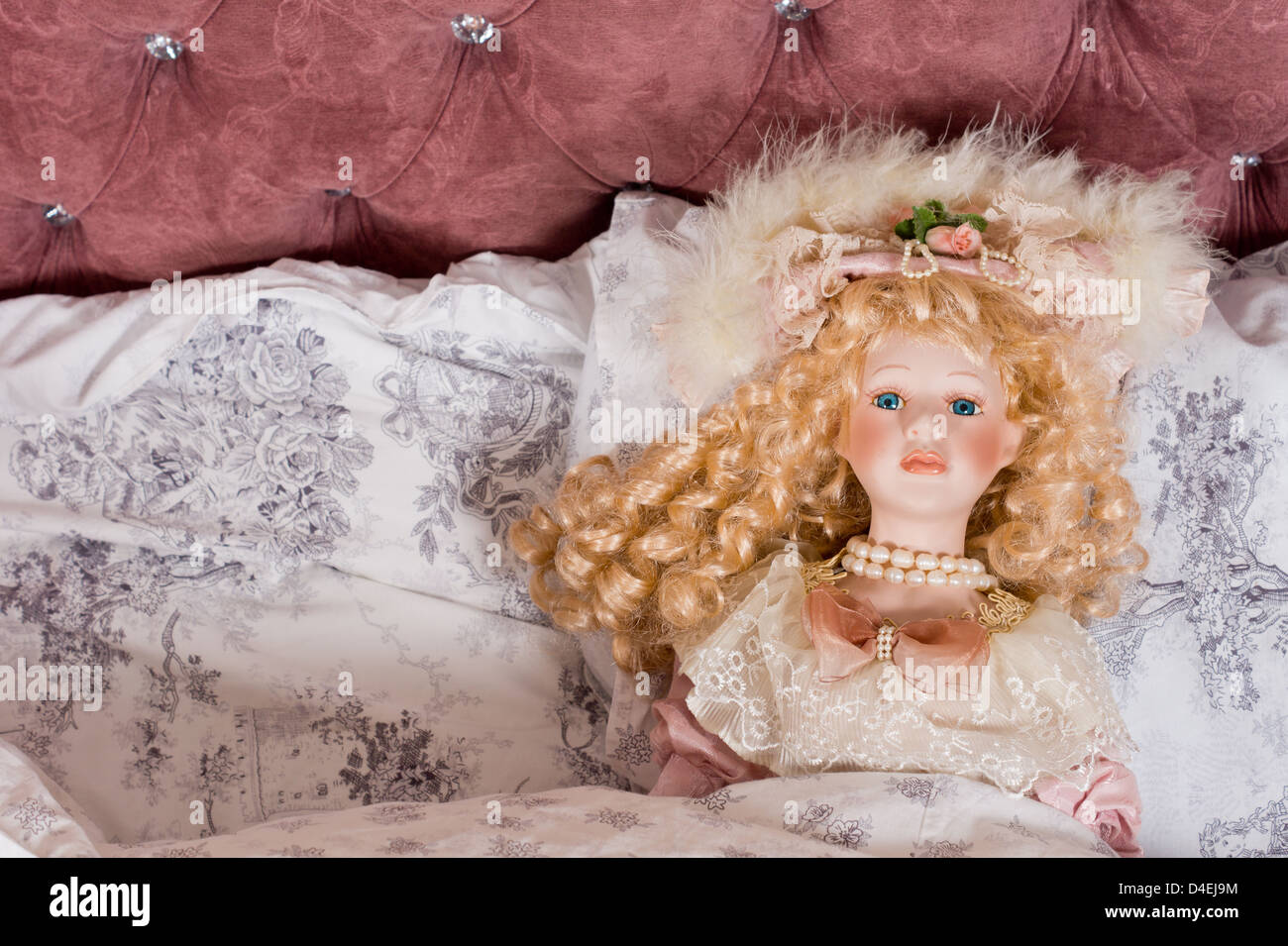 Face of a beautiful vintage doll with her long blonde hair in ringlets and wearing pearls and a fancy hat lying on a bed Stock Photo