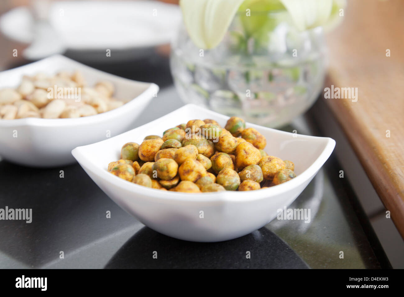 bowl of dehydrated roasted spicy peas in a white square designer porcelain square bowl on table top peanut bowl in background Stock Photo