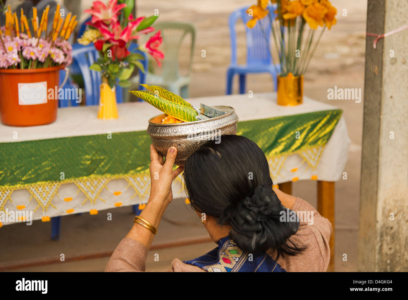 A dark-haired woman holds a metal bowl filled with offerings up to her forehead Stock Photo