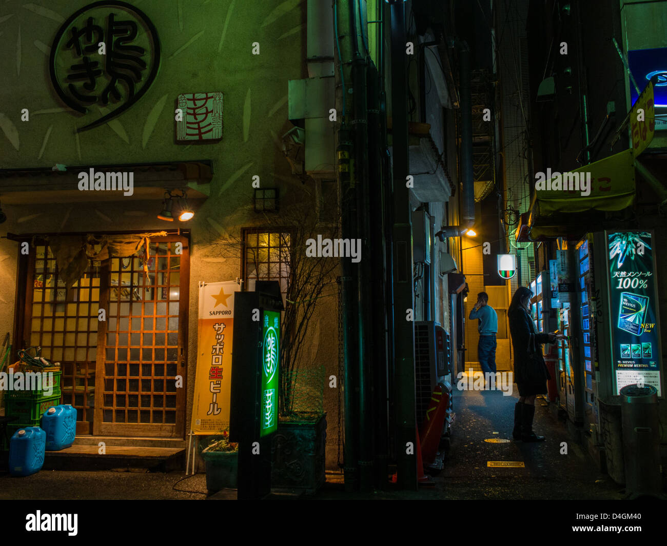 Osaka at night.  In an alley next to a restaurant a woman buys cigarettes from a vending machine while a man talks on his phone. Stock Photo