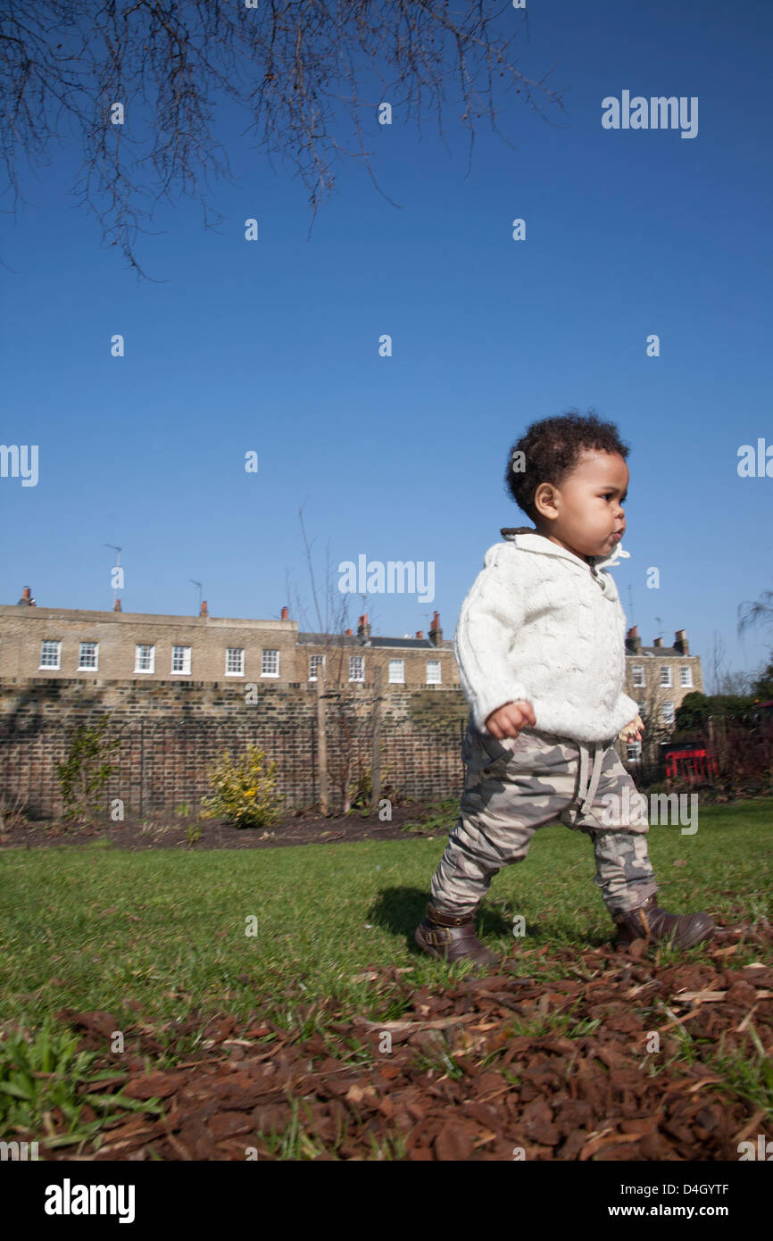 mixed race toddler walking in park wearing cream jumper and combats - green grass and blue sky in background Stock Photo