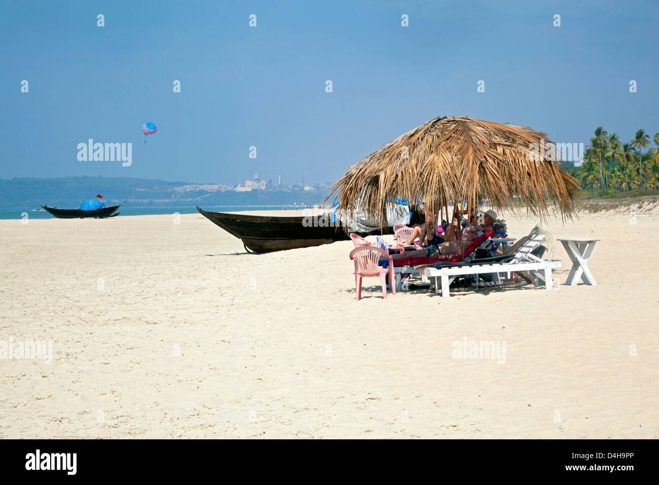 holidaymakers basking in the sun under the shade of coconut parasols. White sand, blue sky, sea, surf and fishing local boats Stock Photo