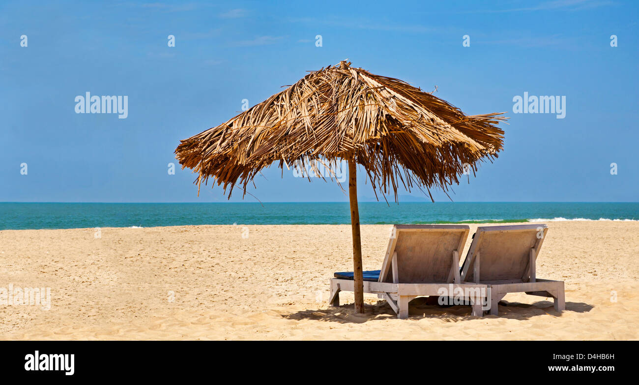 tropical deserted beach with loungers under a coconut leaf parasol. Blue sky, ocean, gentle surf and clean sandy beach. Stock Photo
