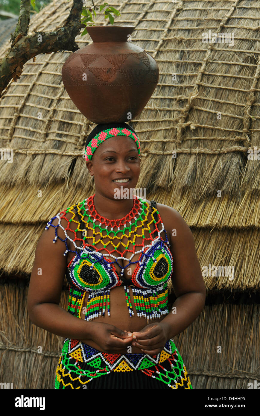 People, Zulu maiden in colourful traditional dress with clay pot on head next to grass hut at tourist attraction Shaka land South Africa Stock Photo