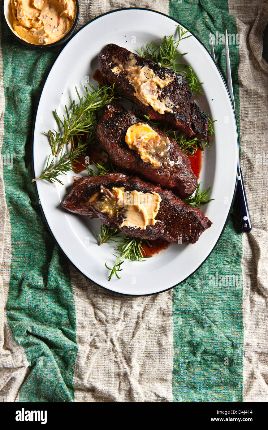 Grilled Denver Steak, topped with Anchovy Butter over Rosemary Sprigs on a white Platter on a striped rustic table cloth. Stock Photo