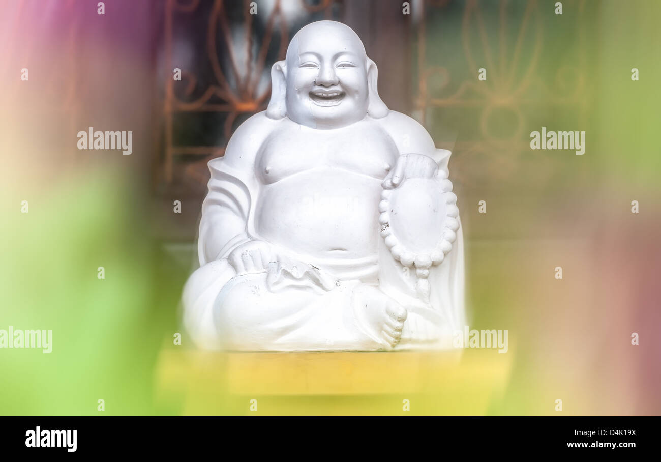 Focus on white statue of laughing monk. Pink and green colors in foreground, window bars in background. Sculpture of fat man sit Stock Photo