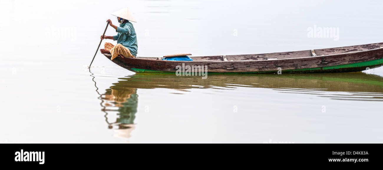 Woman in conical hat sitting on canoe and rowing. Traditional asian boat made of wood. Person holding paddle in hands. Ripple on Stock Photo