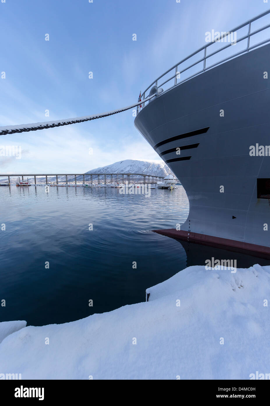A view towards Tromso Bridge with part of a snow-covered wharf and the bows of a moored ship framing the picture. Stock Photo
