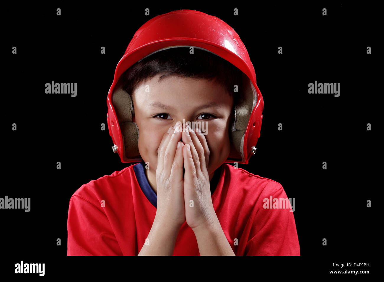 Smiling young boy covers his mouth with hands - with red baseball helmet on black background - dramatic lighting style Stock Photo