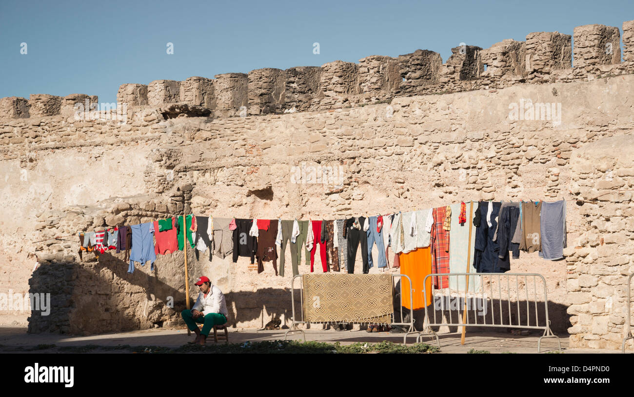 A man sitting next to washing hanging out to dry. Crenelated rampart wall of Essaouira behind. Stock Photo
