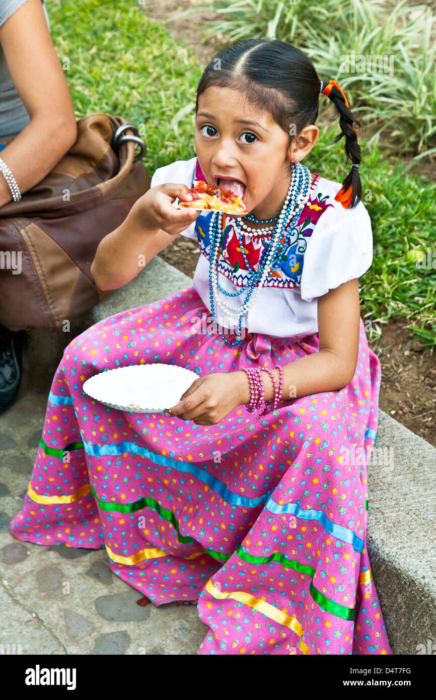 little Mexican girl dressed as Guadalupe in beautifully embroidered skirt blouse eating pizza slice in Llano park Oaxaca Mexico Stock Photo