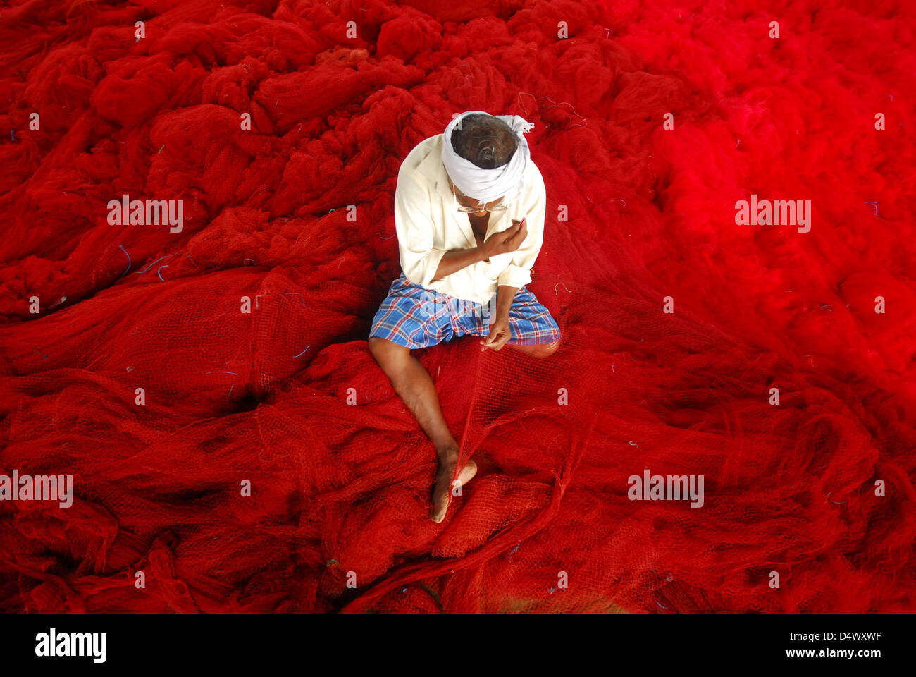 fisherman weaving red coloured fishing net by sitting on that. Stock Photo