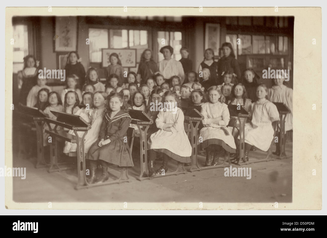 Young Edwardian girls with their arms folded, sitting at desks in a classroom, teachers in the background, at primary school, circa 1908 , U.K. Edwardians Stock Photo
