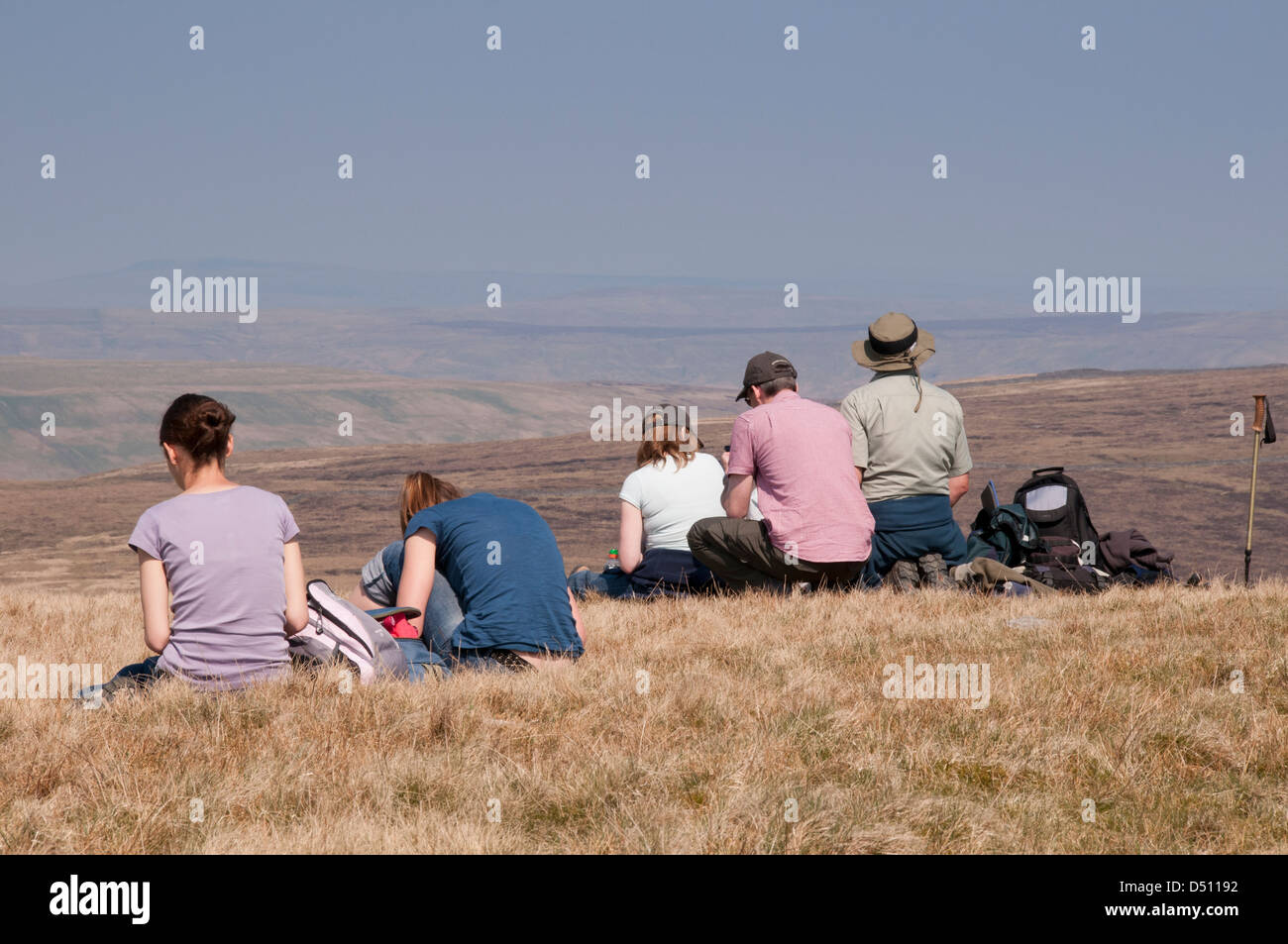 Rear view of people (walkers) sitting on high summit of Pen-y-ghent (one of Yorkshire Three Peaks) looking out over scenic hazy uplands - England, UK. Stock Photo