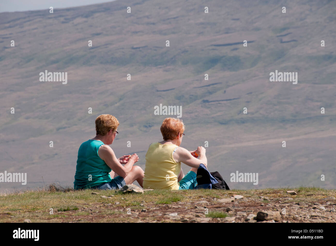 Rear view of 2 women or females (walkers) sitting on sunny summit of Pen-y-ghent, eating picnic, steep fellside beyond - Yorkshire Dales, England, UK. Stock Photo