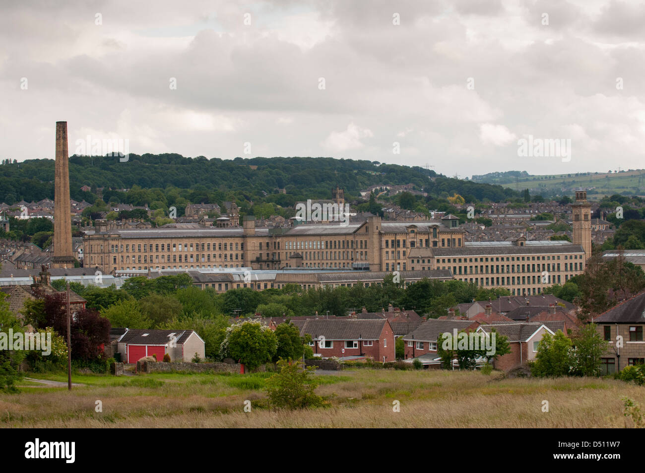 Historic impressive Salts Mill (Victorian textile mill) in Aire valley, tall chimney & weaving sheds towering over houses - Saltaire, England, GB, UK. Stock Photo