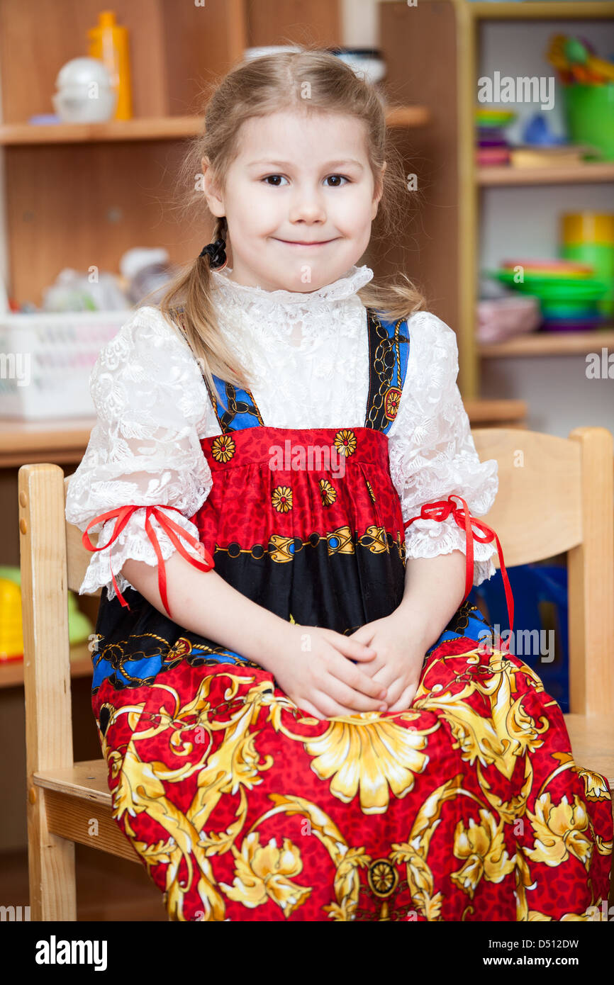Cute girl in Russian traditional clothes in kindergarten sitting on the chairs in classroom