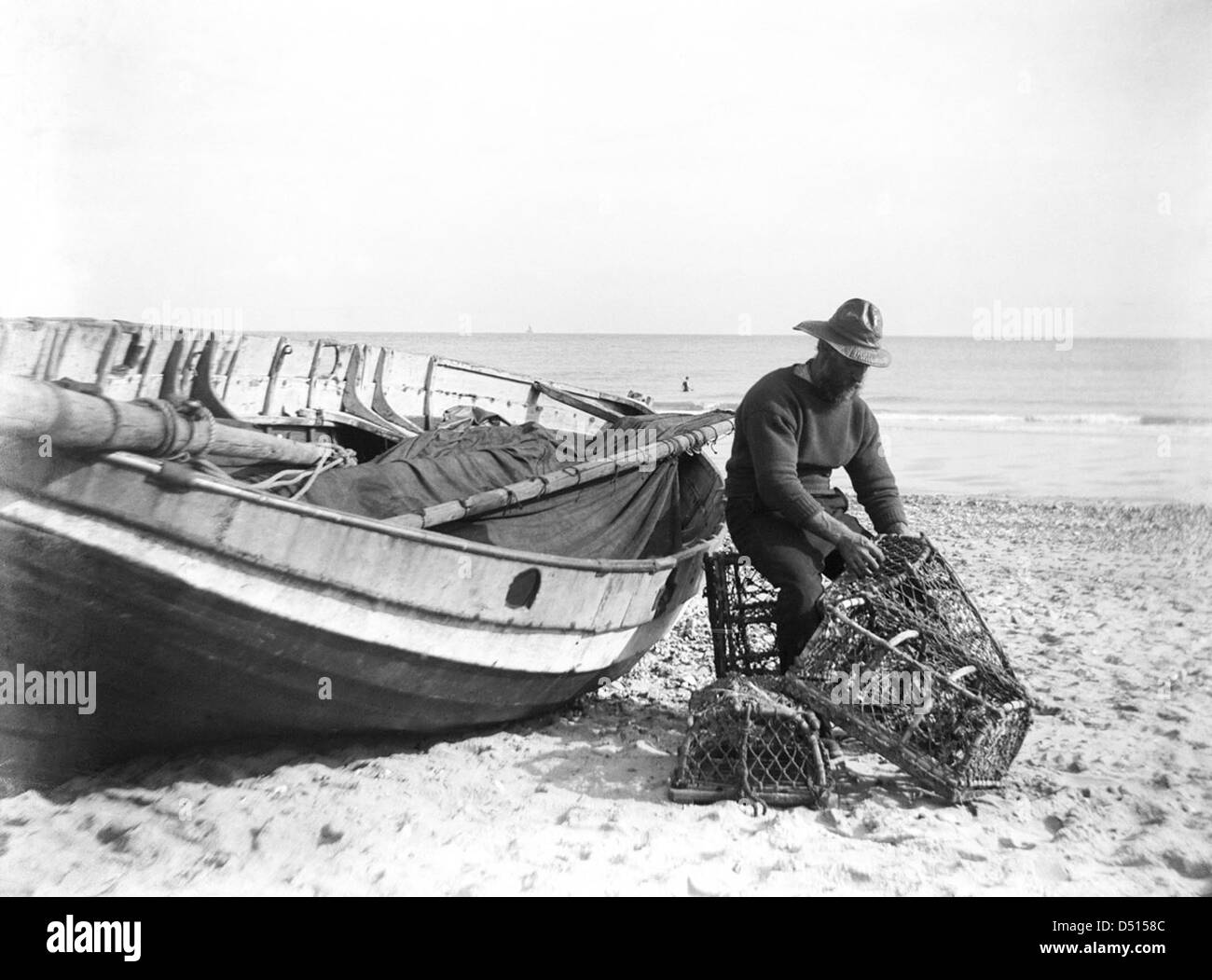 A fisherman in sou'wester mending lobster/crab creels on the beach alongside a beached Sheringham crab boat. Stock Photo