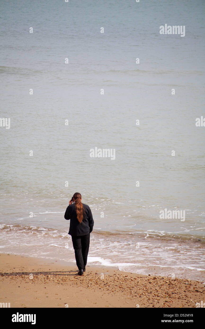 Man with long hair tied in ponytail on mobile phone standing by sea at Bournemouth beach in March Stock Photo