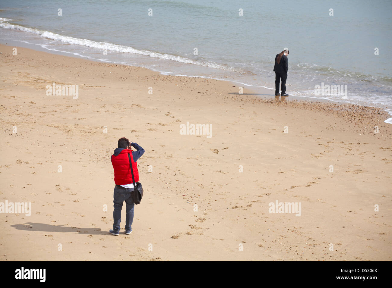 man taking photograph of man with long hair tied in ponytail on mobile phone standing by sea at Bournemouth beach in March Stock Photo